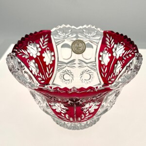 May include: A clear crystal bowl with a ruby red rim and accents. The bowl features a scalloped edge and intricate leaf and floral patterns. A small, circular emblem is visible in the center of the bowl.