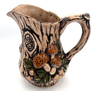 May include: A ceramic pitcher with a tree trunk design. The pitcher is tan with brown bark-like texture and a handle that resembles a tree branch. It features a cluster of orange and white mushrooms with green leaves.