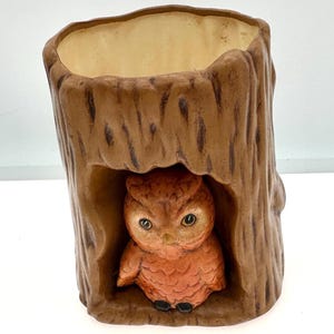 May include: A ceramic planter shaped like a hollow tree trunk with a textured brown bark exterior and a cream-colored interior. An orange owl with black eyes and feet is sculpted into a recessed opening at the front of the tree trunk structure.