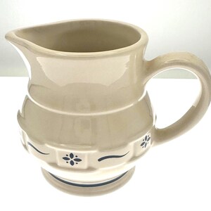 May include: A cream-colored ceramic pitcher featuring a rounded body with a wide handle and a pouring spout. The exterior is decorated with a repeating pattern of dark blue floral motifs and horizontal lines embossed around the lower section.