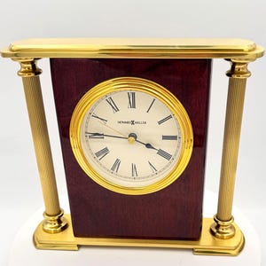 May include: A Howard Miller desk clock with a gold-tone frame and a dark wood finish. The clock face is white with black Roman numerals and gold hands. The clock has a rectangular shape with gold pillars on each side.