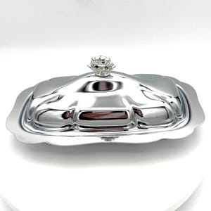 May include: A shiny, silver-colored butter dish with a lid. The lid is topped with a decorative, flower-shaped handle. The dish has a rectangular shape with gently curved edges. The butter dish is on a white surface.