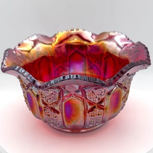 May include: A round, iridescent carnival glass bowl featuring a deep red and purple hue with a metallic sheen. The exterior surface displays a pressed geometric pattern with alternating panels of smooth ovals and textured cross-hatch details.