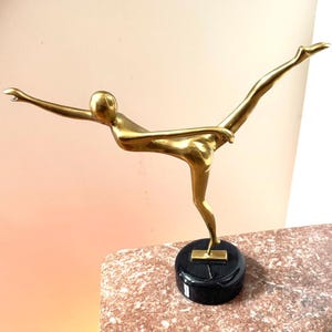 May include: A gold-colored sculpture of a person in a graceful pose, balancing on one leg with arms and the other leg extended. The sculpture is mounted on a black circular base, which sits on a pink and brown marble surface.