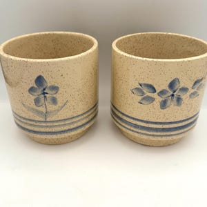May include: Two cylindrical ceramic cups with a cream-colored speckled finish. Each cup features hand-painted blue floral motifs and three horizontal blue stripes encircling the lower portion of the exterior. The cups have a smooth, rounded rim.