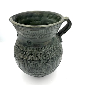 May include: A dark green, antique-style pitcher with a handle and spout. The ceramic vessel features detailed relief carvings of figures and decorative patterns around the body and rim. The pitcher is displayed on a white surface.