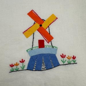 May include: Embroidered white tea towel with a blue and white windmill with orange and yellow blades. There are three red, orange, and yellow flowers with green stems embroidered on the bottom.
