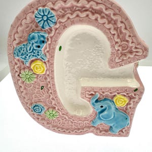 May include: A ceramic letter G in a soft pink hue with a textured lace-like pattern. The surface is decorated with small, raised, colorful figures including a blue lamb, a blue elephant, and several small yellow, green, and blue flowers.