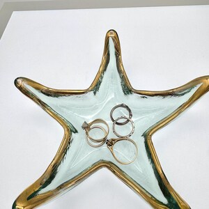 May include: A starfish-shaped glass dish with a pale green center and gold-painted edges. Inside the dish are five metal rings, including gold and silver-toned bands, some featuring small stone settings and textured metal surfaces.