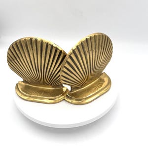 May include: Two gold-colored seashell bookends. Each bookend has a fan-shaped shell design with vertical lines and a curved base. The bookends are made of a shiny, metallic material and are displayed on a white surface.