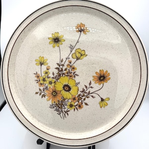 May include: Round ceramic plate with a speckled beige background and a brown rim. The plate features a floral design with yellow and orange flowers and brown stems. A decorative piece for serving food.