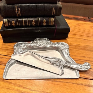 May include: A silver-toned metal crumb tray and matching scraper set featuring ornate, raised floral and foliate relief detailing along the edges and handle. The tray has a wide, flat base, and the scraper has a long, curved, decorative handle.