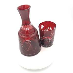 May include: A ruby red glass decanter and tumbler set. The decanter has a long neck and a bulbous base, with etched floral and grape designs. The tumbler is cylindrical, with matching etched details. Both pieces are on a white surface.