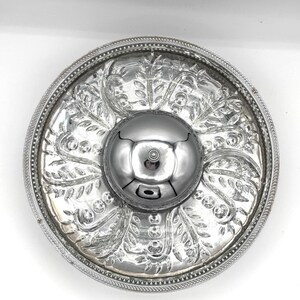 May include: A silver-toned serving dish with a central, raised bowl. The dish features an ornate, embossed floral design and a beaded edge. The bowl reflects the surrounding environment. The overall design is elegant and decorative.