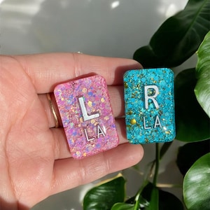 May include: Two small square resin tiles with glitter embedded in the resin. The tiles are pink and blue. The pink tile has a white letter "L" and the text "LA" on it. The blue tile has a white letter "R" and the text "LA" on it.