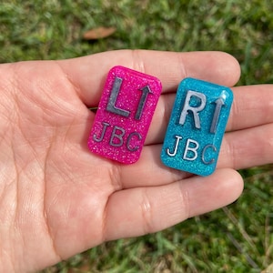 May include: Two small, square-shaped, glittery resin tiles with an arrow pointing upwards. The tile on the left is pink with the letters "L1 JBC" and the tile on the right is blue with the letters "R1 JBC".