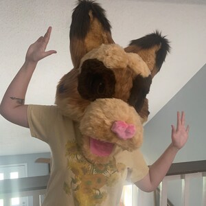 May include: A person wearing a tan furry animal head with brown ears and a pink nose. The head has a large pink tongue sticking out. The person is wearing a yellow t-shirt with a sunflower print.