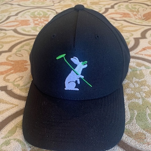 Peut inclure: Casquette de baseball noire avec un lapin blanc tenant un club de golf vert brodé sur le devant.