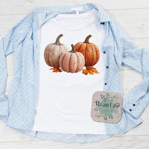 Op de afbeelding: Wit T-shirt met een ontwerp van drie pompoenen in beige, perzik en oranje, met herfstbladeren. Een lichtblauw overhemd met knopen is eroverheen gelegd. Een onderzetter met de tekst "Ma'am Cave Creations" is ook zichtbaar.