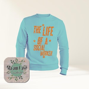Peut inclure: Un sweat-shirt bleu clair avec le texte "THE LIFE OF A SOCIAL WORKER" en paillettes orange. Le sweat-shirt a des manches longues et un col rond. Un dessous de verre en bois avec le texte "Ma'am Cave Creations" est dans le coin inférieur gauche.