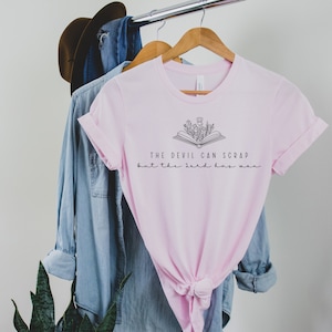 May include: Pink t-shirt with a black graphic of an open book with flowers growing out of it. The text "The devil can scrap but the Lord has won" is printed below the book.