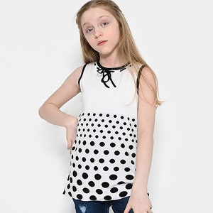 May include: A white sleeveless top with a black trim and a black polka dot pattern on the bottom half. The top has a black tie at the neckline.