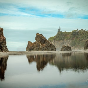 May include: A scenic view of a rocky coastline with three tall rock formations rising from the water. The rocks are reflected in the calm water, creating a mirror-like effect. The sky is a light blue with wispy clouds.
