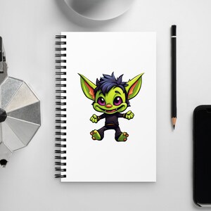 Puede incluir: Un cuaderno blanco con espiral con una ilustración de un duende de dibujos animados. El duende tiene piel verde, ojos morados y ropa negra. También se ven una taza de café, un lápiz, un teléfono y una cafetera plateada.