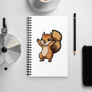 May include: A white spiral-bound notebook featuring a cartoon squirrel illustration. The squirrel is brown and tan with a fluffy tail and is drawn with its arms raised. A cup of coffee, a pencil, a phone, and a coffee maker are also on the white surface.