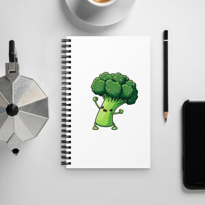 May include: White spiral-bound notebook with a cartoon broccoli character with a smiling face and raised arms. The notebook is on a white surface, next to a cup of coffee, a pencil, a smartphone, and a silver coffee maker.