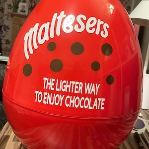 Puede incluir: Un gran recipiente rojo en forma de huevo con la palabra "Maltesers" en escritura blanca. El recipiente tiene acentos circulares marrones y el texto "THE LIGHTER WAY TO ENJOY CHOCOLATE" en blanco.