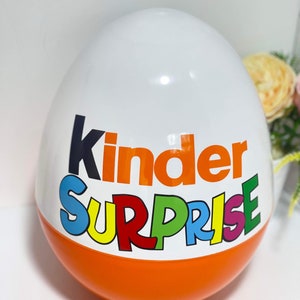 Peut inclure: Un œuf en plastique blanc avec les mots "Kinder Surprise" imprimés en orange et en arc-en-ciel. L'œuf est posé sur une surface blanche.