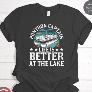 May include: A dark heather grey t-shirt with a vintage-style graphic of a pontoon boat on a lake. The text "Pontoon Captain" is in a white circle above the boat, and the text "Life is Better at the Lake" is below the boat.