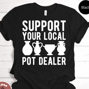 May include: Black t-shirt with the white text "SUPPORT YOUR LOCAL POT DEALER" and four white pottery illustrations. The shirt is made of a soft material and is suitable for casual wear.