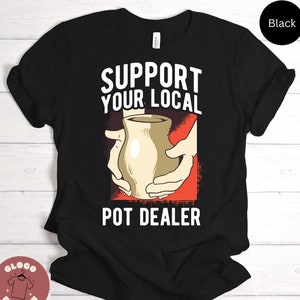 May include: Black t-shirt with a white graphic that says "Support Your Local Pot Dealer". The graphic features a hand holding a clay pot.