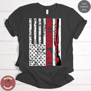 May include: A dark heather grey t-shirt with a distressed American flag design and the text "TRAP SHOOTING" in red. A red shotgun is depicted on the right side of the flag.
