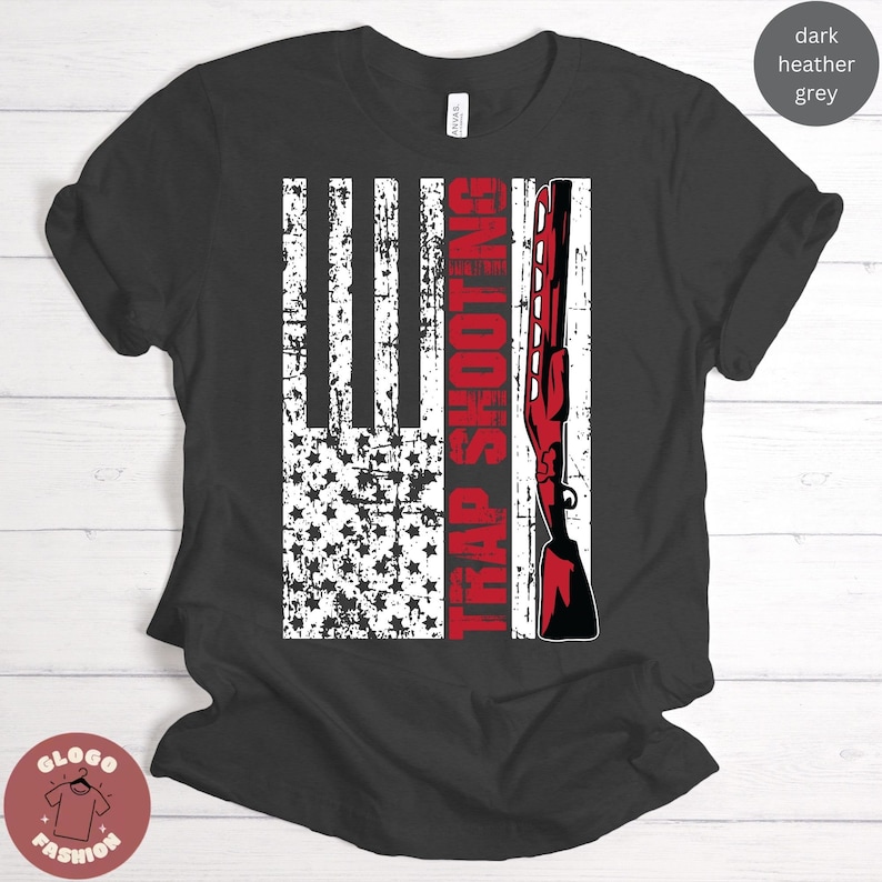 May include: A dark heather grey t-shirt with a distressed American flag design and the text "TRAP SHOOTING" in red. A red shotgun is depicted on the right side of the flag.