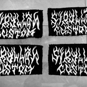 Op de afbeelding: Vier zwarte rechthoekige patches met witte, gestileerde tekst. De tekst lijkt een bandnaam of logo te zijn. De patches zijn in een vierkant gerangschikt, twee boven en twee beneden.