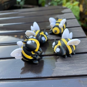May include: Four 3D printed bumblebees in black and yellow with white wings. The bumblebees are sitting on a dark wooden surface.