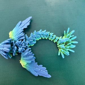 May include: A 3D printed blue and green dragon figurine with a long, spiky tail and wings. The dragon is made of plastic and has a textured surface.