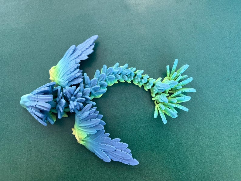 May include: A 3D printed blue and green dragon figurine with a long, spiky tail and wings. The dragon is made of plastic and has a textured surface.