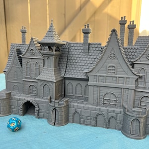 May include: A gray 3D printed model of a large, multi-sectioned gothic style house with a tower and multiple chimneys. The house is sitting on a blue surface with a blue and gold dice in front of it.