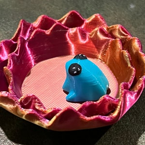 May include: A small, blue frog figurine sits inside a pink and gold, 3D printed, flower-shaped dish.