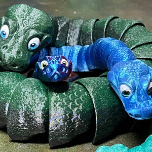 Puede incluir: Un grupo de tres juguetes de serpiente segmentados, de color verde, azul y turquesa. Las serpientes est&aacute;n enroscadas juntas, con las cabezas de cada serpiente visibles. Las serpientes tienen ojos grandes y redondos y una superficie texturizada.