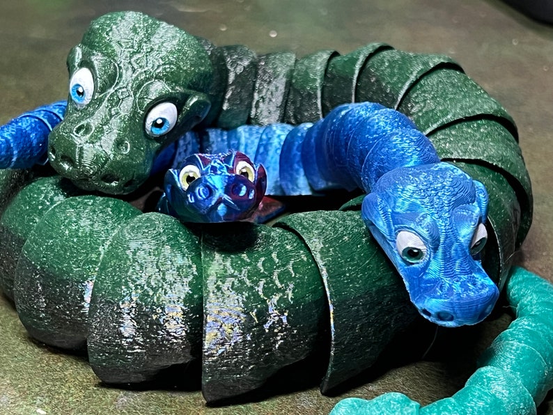 Puede incluir: Un grupo de tres juguetes de serpiente segmentados, de color verde, azul y turquesa. Las serpientes est&aacute;n enroscadas juntas, con las cabezas de cada serpiente visibles. Las serpientes tienen ojos grandes y redondos y una superficie texturizada.