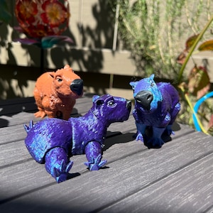 May include: Three toy capybaras, one brown and two purple, are posed on a gray surface. The purple capybaras have a shimmery, iridescent finish.