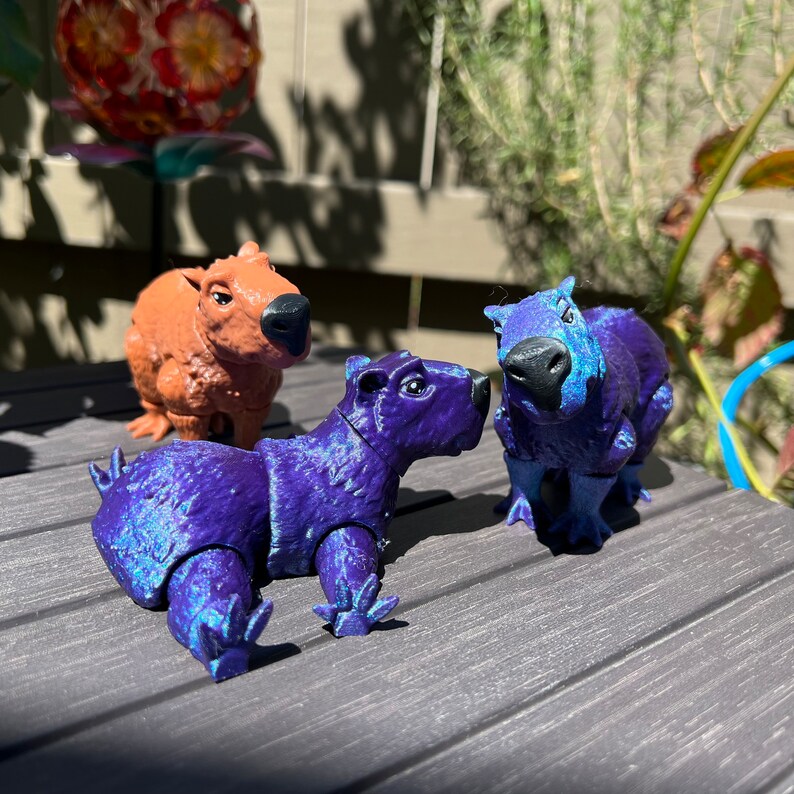 May include: Three toy capybaras, one brown and two purple, are posed on a gray surface. The purple capybaras have a shimmery, iridescent finish.