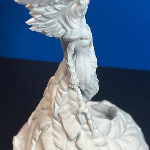 Puede incluir: Escultura blanca detallada de una figura alada, posiblemente un ángel o criatura mítica. La figura tiene cabello suelto y está sobre una base texturizada. Las alas están extendidas y el diseño es intrincado.