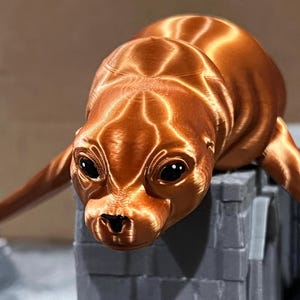 May include: A 3D printed figurine of a seal, with a brown, shiny finish. The seal is lying on its belly with its head turned to the side, looking at the camera. The seal's eyes are black and its nose is brown.