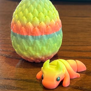 May include: A small, colorful dragon figurine with a gradient of yellow, orange, and red. The dragon is curled up in a resting position. It is next to a larger, egg-shaped object with a similar color gradient. The egg is covered in small, raised scales.